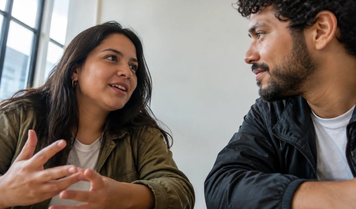 Três participantes conversam ao redor de uma mesa comprida numa sala clara de janelas altas